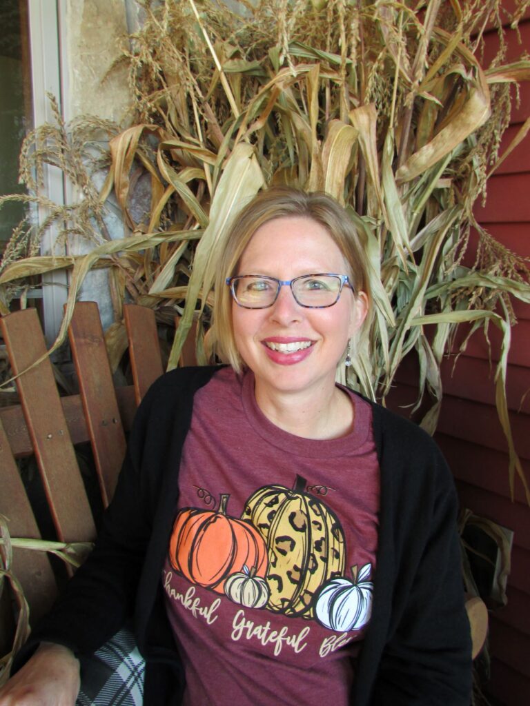 woman sitting with cornstalks in background with a shirt with pumpkins and thankful grateful blessed on her shirt