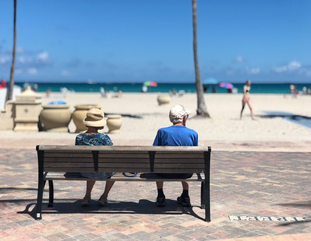 2 people sitting on bench watching people by the beach